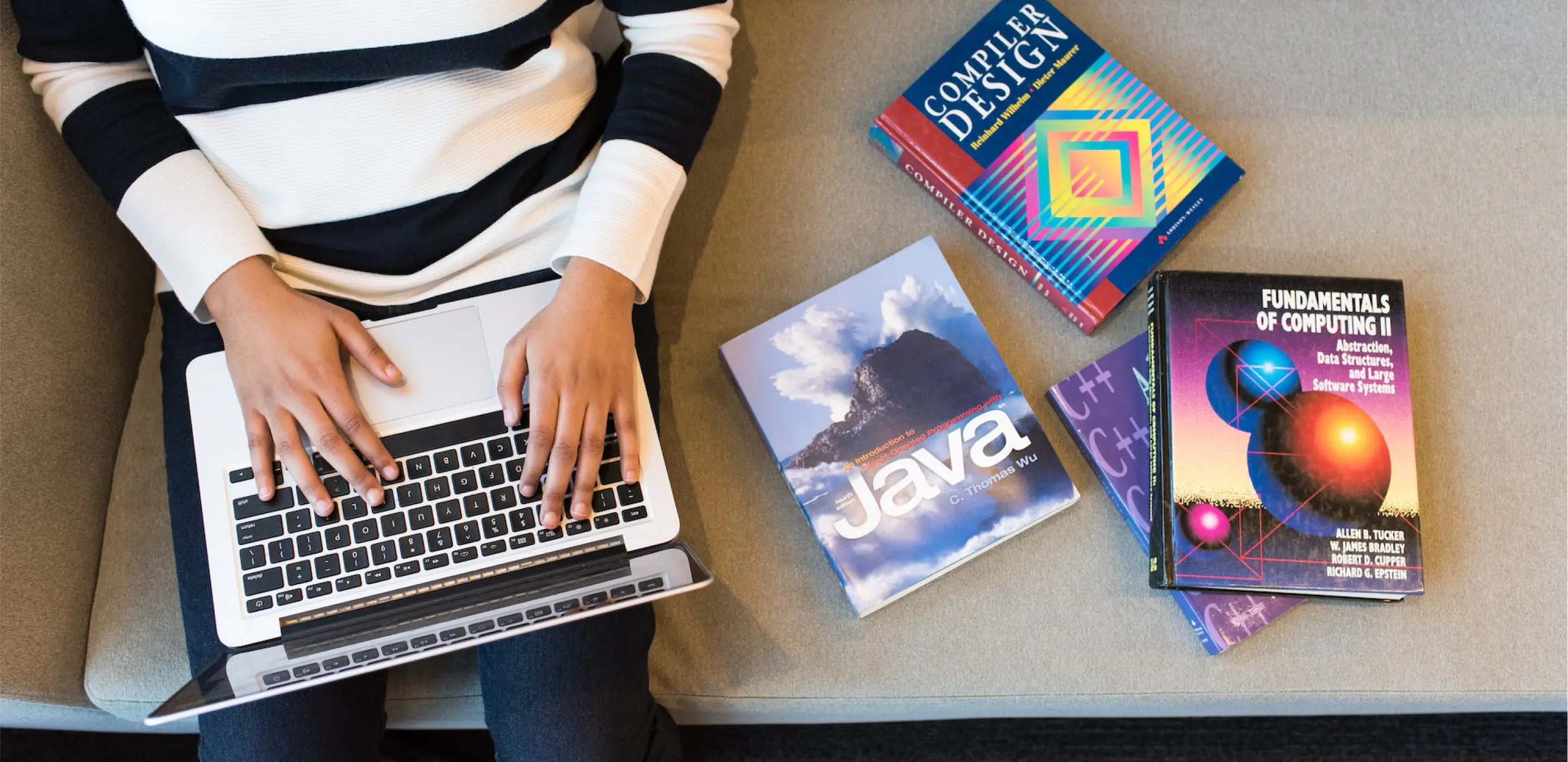 Seseorang mengetik di laptop mereka di sofa, dengan buku yang tersebar di samping